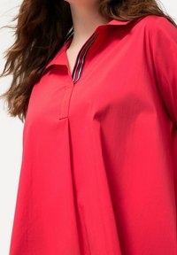 Punainen paita, jossa on kaulus ja raidallinen pääntie. Pehmeä kangas ja väljä malli, jossa on hieno poimutuskaulus. 