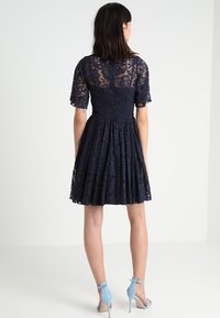 Marinblå spetsklänning med korta ärmar, figursydd överdel och utsvängd kjol. Har blommönster och en nyckelhålbackdetalj.