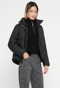 Svart foret jakke med hette, glidelås og trykknapper, kombinert med en svart genser med høy hals og bukser med grå leopardmønster.