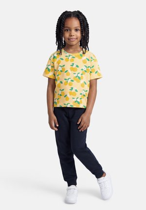 Niña joven con el cabello trenzado, vistiendo una camisa amarilla con estampado de frutas, pantalones azul marino y zapatillas blancas, de pie contra un fondo liso.