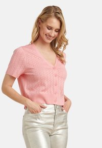 Kurzärmlige rosa Strickjacke mit V-Ausschnitt, die ein strukturiertes Diamantmuster und sechs Knöpfe aufweist, kombiniert mit metallisch silbernen Hosen.