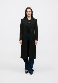 Hosszú fekete kabát szabott kialakítással, egysoros gombolással és hajtókás gallérral, fekete inggel és sötétkék farmernadrággal párosítva.