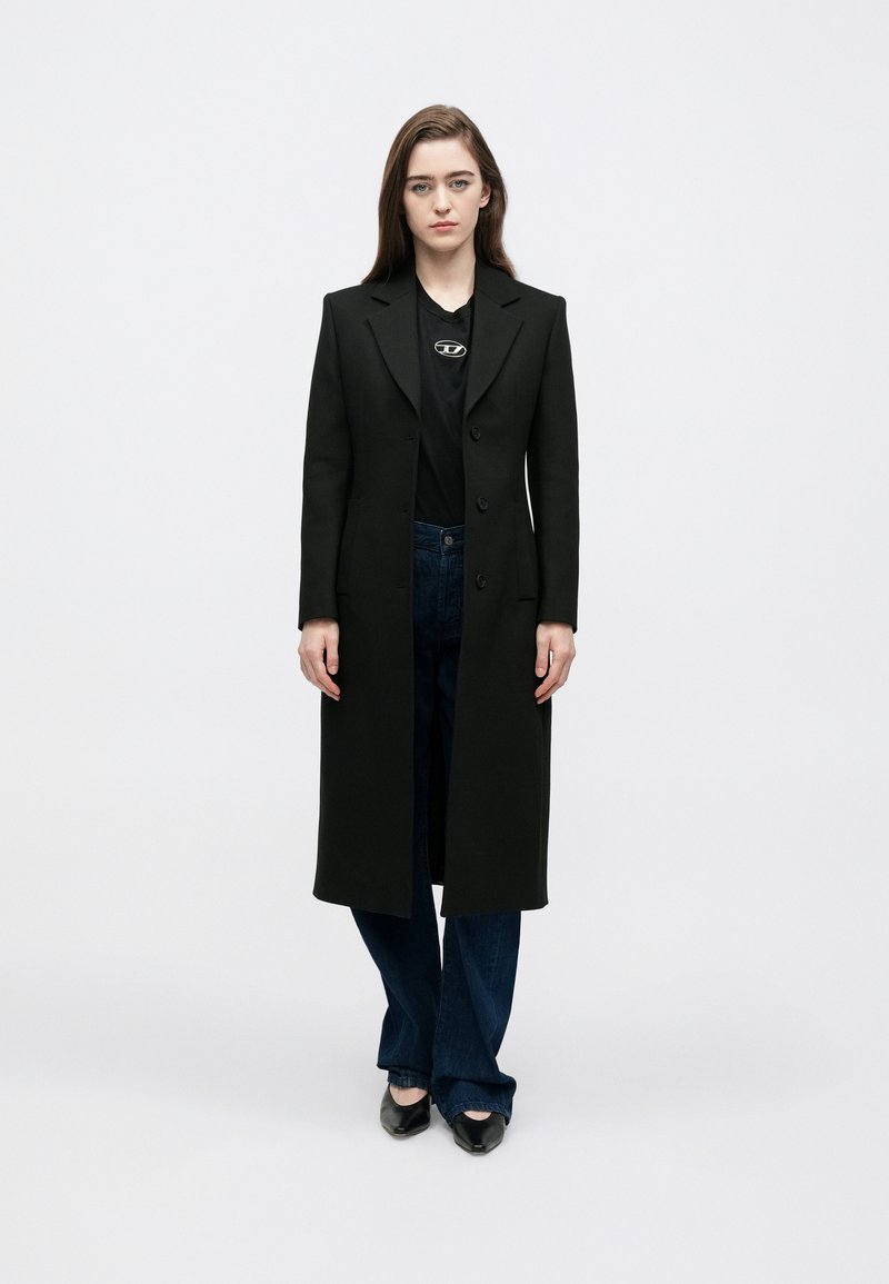 Hosszú fekete kabát szabott kialakítással, egysoros gombolással és hajtókás gallérral, fekete inggel és sötétkék farmernadrággal párosítva.