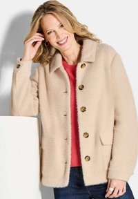 Licht beige gestructureerde jas met knopen, een kraag en twee voorzakken, gedragen over een rode top en donkere jeans.