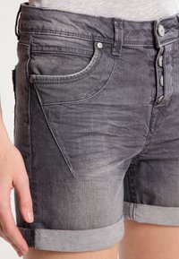 Harmaat farkkushortsit, joissa haalistettu pinta, viiden taskun malli ja käännetyt reunat. Varustettu metallisilla yksityiskohdilla ja tikatuilla koristeilla.