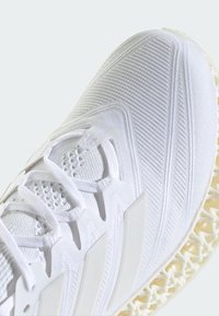White athletic shoe with a knitted upper, textured detailing, and a distinctive patterned sole. Features white laces and branding accents.