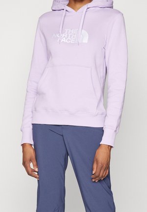 Osoba ubrana w jasnofioletową bluzę z kapturem The North Face i ciemnoniebieskie spodnie, stojąca z rozluźnionymi rękami wzdłuż boków.