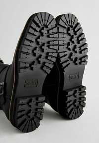 Bottes en cuir noir avec une semelle en caoutchouc texturé présentant de profonds motifs de crampons, accentuées par une boucle décorative et une fermeture éclair.