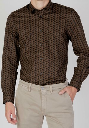 Mann trägt ein braun gemustertes, langärmliges Hemd mit Knöpfen, das in beige Hosen gesteckt ist, mit einer Hand in der Hosentasche.