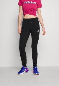 Černé teplákové kalhoty s bílými bočními pruhy a logem Adidas. Kombinované s růžovým krátkým tričkem a modrými sportovními botami.