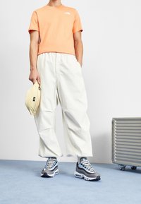 Pomarańczowa koszulka z krótkim rękawem, białe luźne spodnie, szaro-niebieskie wzorzyste trampki i beżowa torba w ręku.