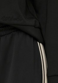 Juodas sportinis kostiumas, pagamintas iš minkštos medžiagos, su iškilniu "Attitude" logotipu ir kontrastingomis šoninėmis juostelėmis smėlio ir rudos spalvos.