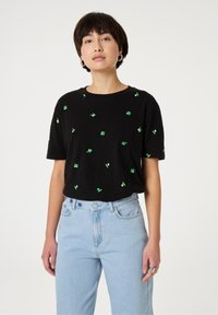 Zwart katoenen T-shirt met korte mouwen, versierd met verspreide groene bladvormige borduursels. Gecombineerd met lichtblauwe high-waisted jeans.