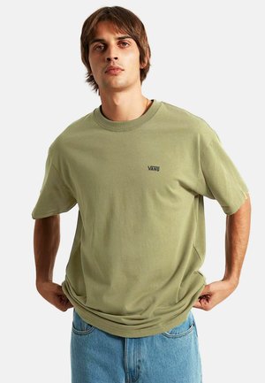 Joven con camiseta Vans verde claro y vaqueros azules, ajustando el bajo de la camiseta, mirando hacia adelante sobre un fondo liso.