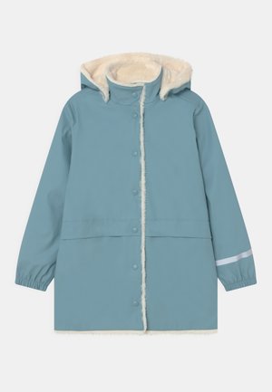 Chaqueta impermeable azul con capucha forrada de piel sintética, cierres de presión, puños elásticos y una franja reflectante en la manga. Textura suave.