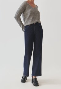 Kobieta ubrana w szary sweter robiony na drutach, ciemnoniebieskie dżinsy z wysokim stanem i czarne loafersy na platformie, stoi z rękami złożonymi za plecami.