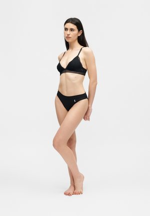 Tânără desculță purtând un bralette de dantelă neagră și chiloți asortați negri cu logo, pe un fundal simplu deschis la culoare.