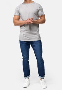 T-shirt gris à manches courtes avec un col rond, associé à un jean en denim bleu et des baskets blanches. Le t-shirt présente un petit logo sur la poitrine.