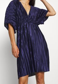 Lila veckad klänning med V-ringning, korta vida ärmar och lös passform. Tyget har en slät, glänsande yta och vertikala ränder.