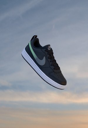 Μαύρο αθλητικό παπούτσι Nike με λευκή σόλα και γκρι σήμα, να αιωρείται πάνω από ένα μαλακό, συννεφιασμένο φόντο ουρανού.
