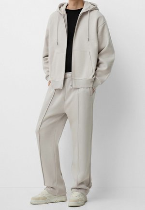 Mann trägt eine hellbeige Kapuzenjacke mit Reißverschluss, dazu passende locker sitzende Jogginghosen, ein schwarzes Shirt und cremefarbene Sneakers, steht mit den Händen in den Taschen.