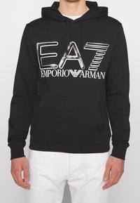 Sudadera negra con gran logo blanco de EA7 Emporio Armani en el pecho, llevada sobre una camisa blanca, combinada con pantalones blancos.