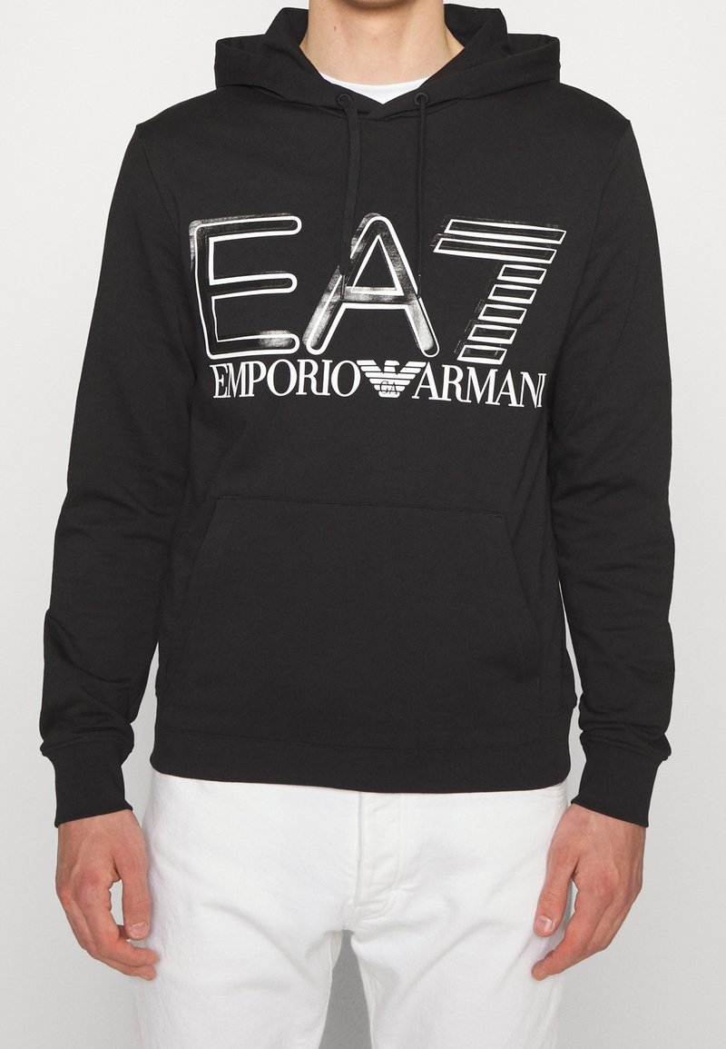Sudadera negra con gran logo blanco de EA7 Emporio Armani en el pecho, llevada sobre una camisa blanca, combinada con pantalones blancos.