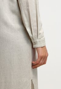 Chemise en lin gris clair à manches longues, dotée d'un poignet à bouton et d'une fente latérale, mettant en valeur la texture naturelle du tissu.