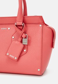 Sac à main en cuir corail avec une forme structurée, accents en métal argenté, logo de la marque embossé et une étiquette détachable attachée par un cadenas.