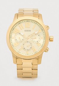 Relógio de pulso Guess em tom dourado com pulseira de metal em elos, mostrador grande e redondo, três submostradores e marcadores de horas dourados sobre um fundo creme.