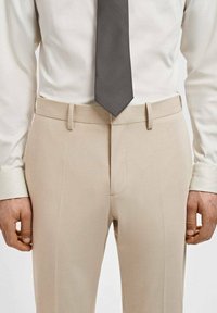 Bež hlače s ravnim prednjem delu, opremljene s pasom, stranskimi žepi in ozkim krojem. V kombinaciji z svetlo obarvano srajco in temnim kravato.