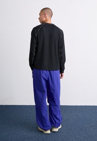Camisa negra de manga larga con costuras grises, combinada con pantalones sueltos de longitud hasta el tobillo en color púrpura, sobre un fondo liso.