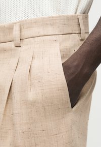 Main à peau foncée placée à l'intérieur de la poche d'un pantalon plissé à taille haute beige avec passants de ceinture.