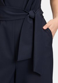 Marinblå jumpsuit med bälte i midjan. Har en mjuk textur och sidofickor. Det mjuka tyget faller elegant.