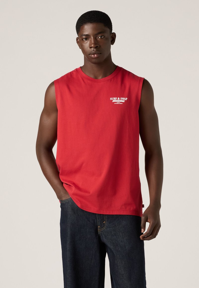 Junger Mann trägt rotes ärmelloses Shirt mit weißem Text und dunkelblauen Jeans, steht vor einem einfarbigen hellen Hintergrund.