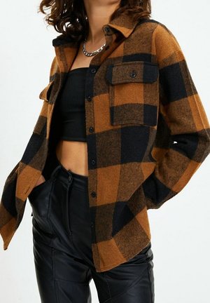 Vrouw met een bruin-zwart geruit jasje met knopen over een zwart cropped topje en een zwarte high-waisted leren broek met kettingketting.