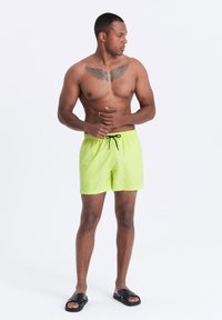 Neonvihreät uima-asut narulla, kevyttä materiaalia ja sivutaskuja. Mustat sandaalit viimeistelevät ilmeen. Iho näkyy vyötärön yläpuolella.