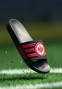 Μαύρη σαγιονάρα Adidas με κόκκινες λεπτομέρειες, λογότυπο και ρίγες. Κατασκευασμένη από λείο υλικό με επίπεδη σόλα, τοποθετημένη πάνω σε φόντο με γρασίδι.