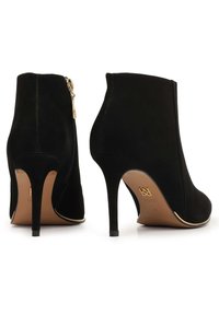 Para czarnych zamszowych botków za kostkę z wysokimi szpilkami i złotym suwakiem z boku, pokazanych z tyłu i z boku na białym tle.