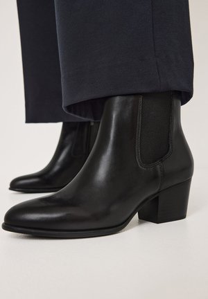 Botas de cuero negras hasta el tobillo con puntera puntiaguda, paneles laterales elásticos y un tacón grueso. Superficie texturizada y detalles de diseño mínimos.