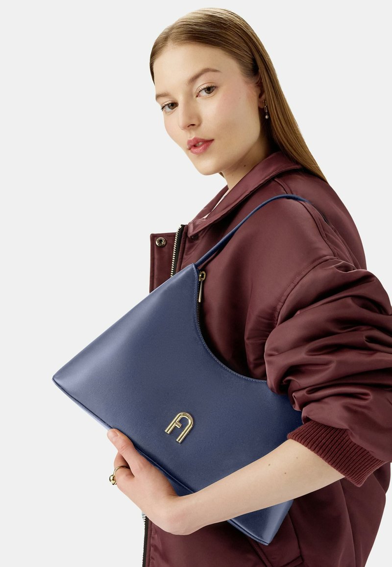 Granatowa skórzana torebka o gładkiej fakturze, zaokrąglonym dnie, złotych akcentach i zamknięciu na zamek, trzymana na tle pluszowej burgundowej kurtki.
