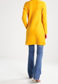 Dlouhý žlutý tunikový svetr s přímým střihem, dlouhými rukávy, nošený přes rozšířené modré džíny. Hladká pletená textura, minimalistické detaily.