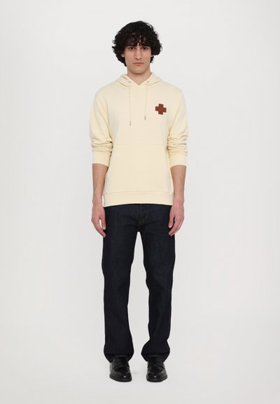 Cream hoodie with a brown cross patch, drawstrings, and kangaroo pocket paired with dark denim jeans and black shoes.