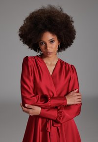 Robe en satin rouge à manches longues, présentant un design croisé et des épaules froncées. Accentuée par une taille nouée et des détails en bouton aux poignets.