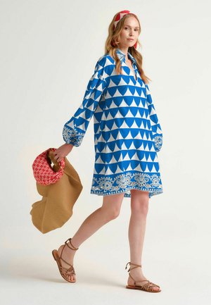 Donna che indossa un vestito azzurro e bianco con motivo, fascia rossa e bianca, porta un cappello intrecciato beige e una borsa rossa con motivo, cammina con sandali.