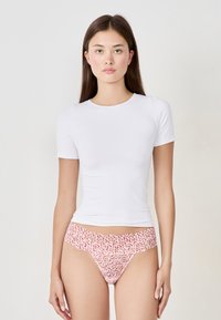 Jeune femme aux longs cheveux bruns, portant un t-shirt blanc à manches courtes et un soutien-gorge en dentelle rose avec des motifs de cœurs rouges, se tenant devant un fond uni.
