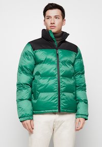 Grüne Pufferjacke mit einem schwarzen oberen Teil, aus glänzender Nylon gefertigt. Verfügt über einen Reißverschluss auf der Vorderseite, einen hohen Kragen und elastische Bündchen.