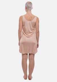 Persona con capelli corti chiari sta a piedi nudi rivolta verso l'elemento opposto, indossando un vestito senza maniche, lungo fino al ginocchio, di un leggero colore pesca, su uno sfondo bianco semplice.