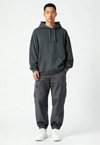 Felpa con cappuccio grigio scuro con tasca a marsupio frontale, abbinata a pantaloni cargo grigio scuro. Sneakers bianche completano l'outfit.