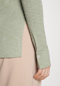 Ljusgrön ribbad tröja med sidoklyftor och knäppte manschetter, parat med en mjuk beige tyg under. Texturerad stickad design är synlig.